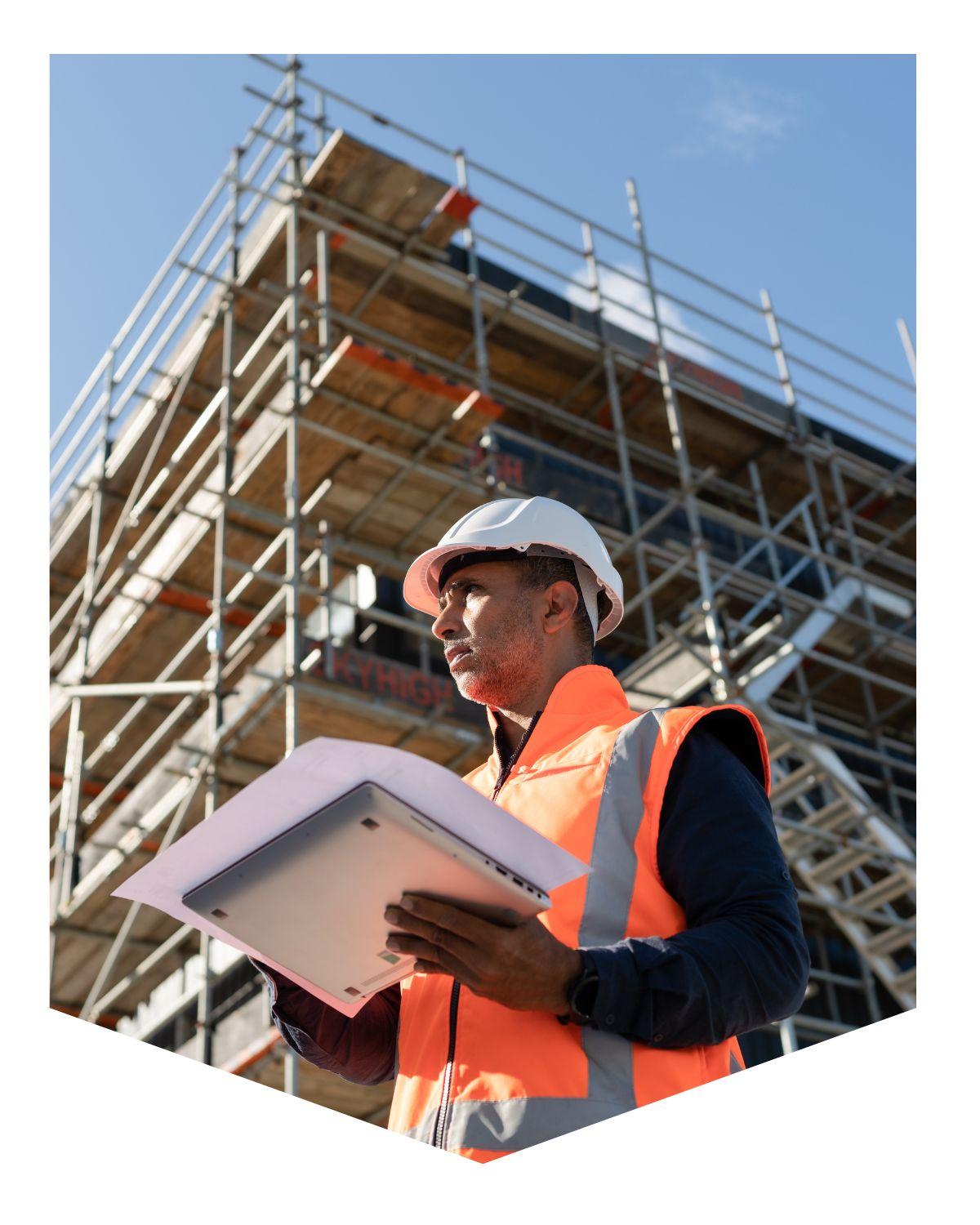 Construction worker standing in front of a building under construction holding a laptop with paper on it