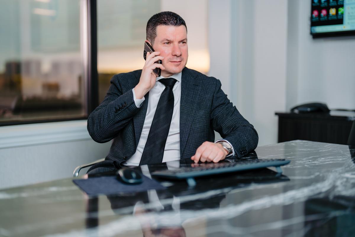 Sam Faris in a dark plaid suit and black tie talking on a smartphone at a marble office desk with a wireless keyboard and mouse in front of him. A large window and wall-mounted screen are visible in the background.