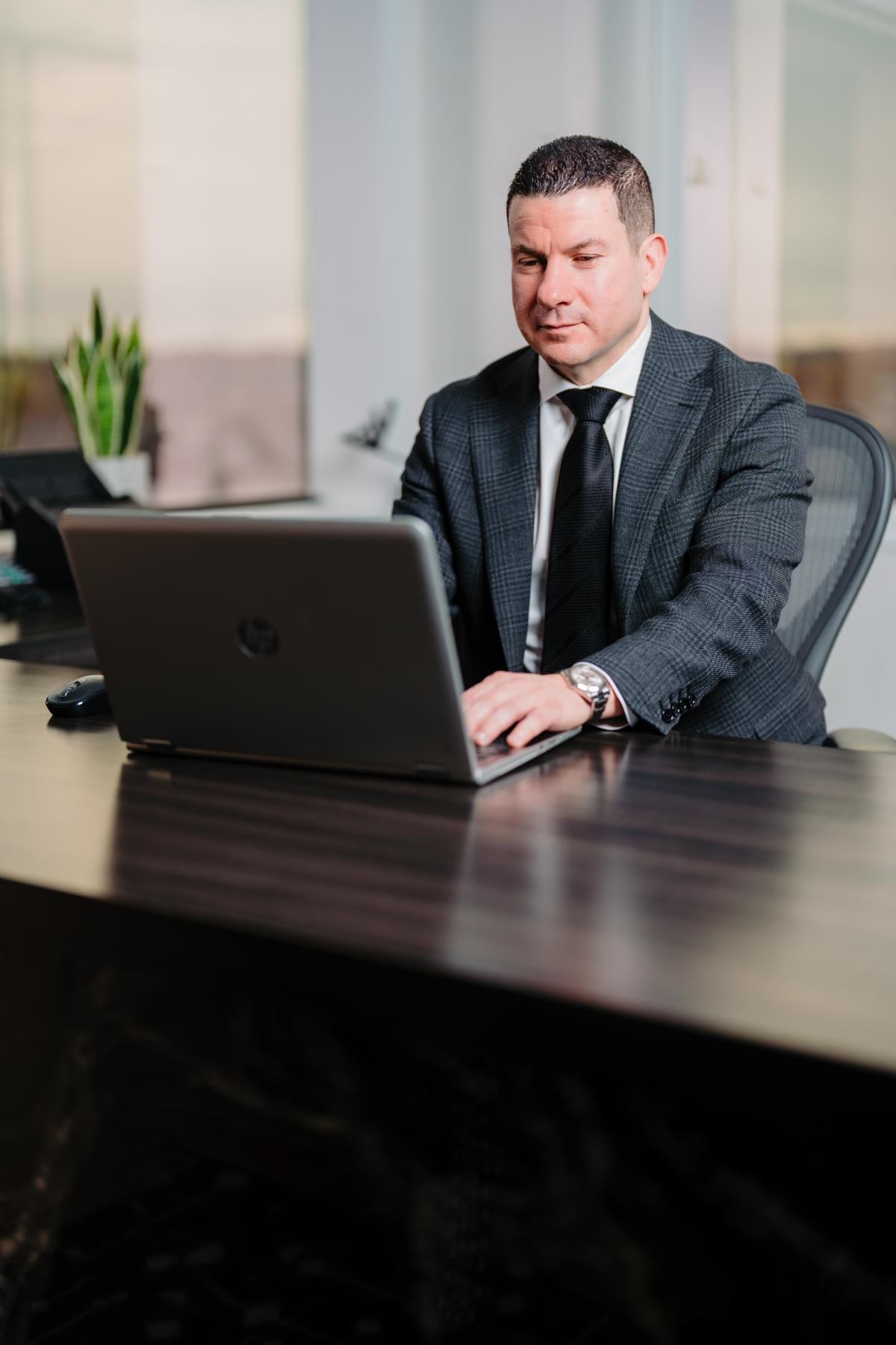 Sam Faris in a business suit working on a laptop at a modern office desk with a plant and blurred cityscape in the background.