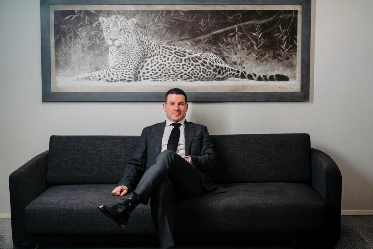 Sam Faris sitting on a couch in a business suit with legs crossed looking at the camera.