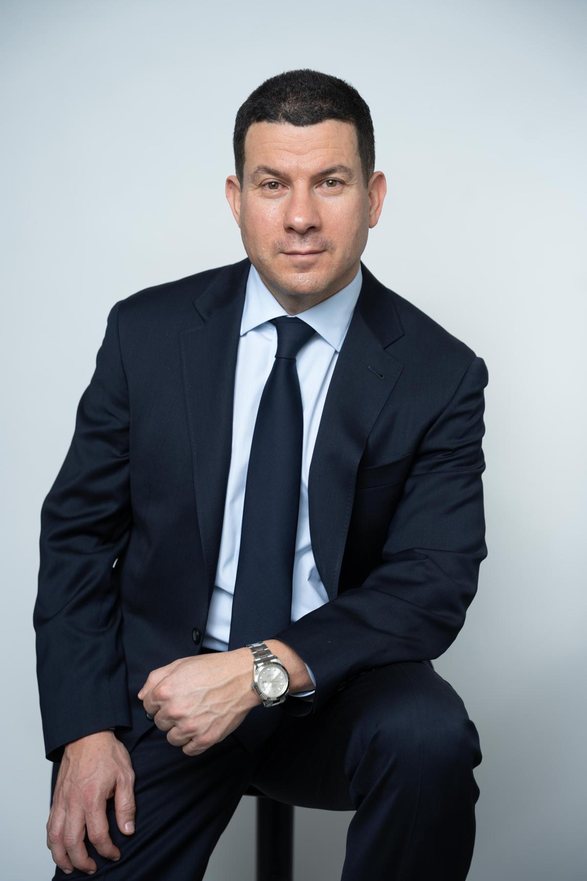Sam wearing a blue suit and sitting on a chair smiling and looking at the camera.