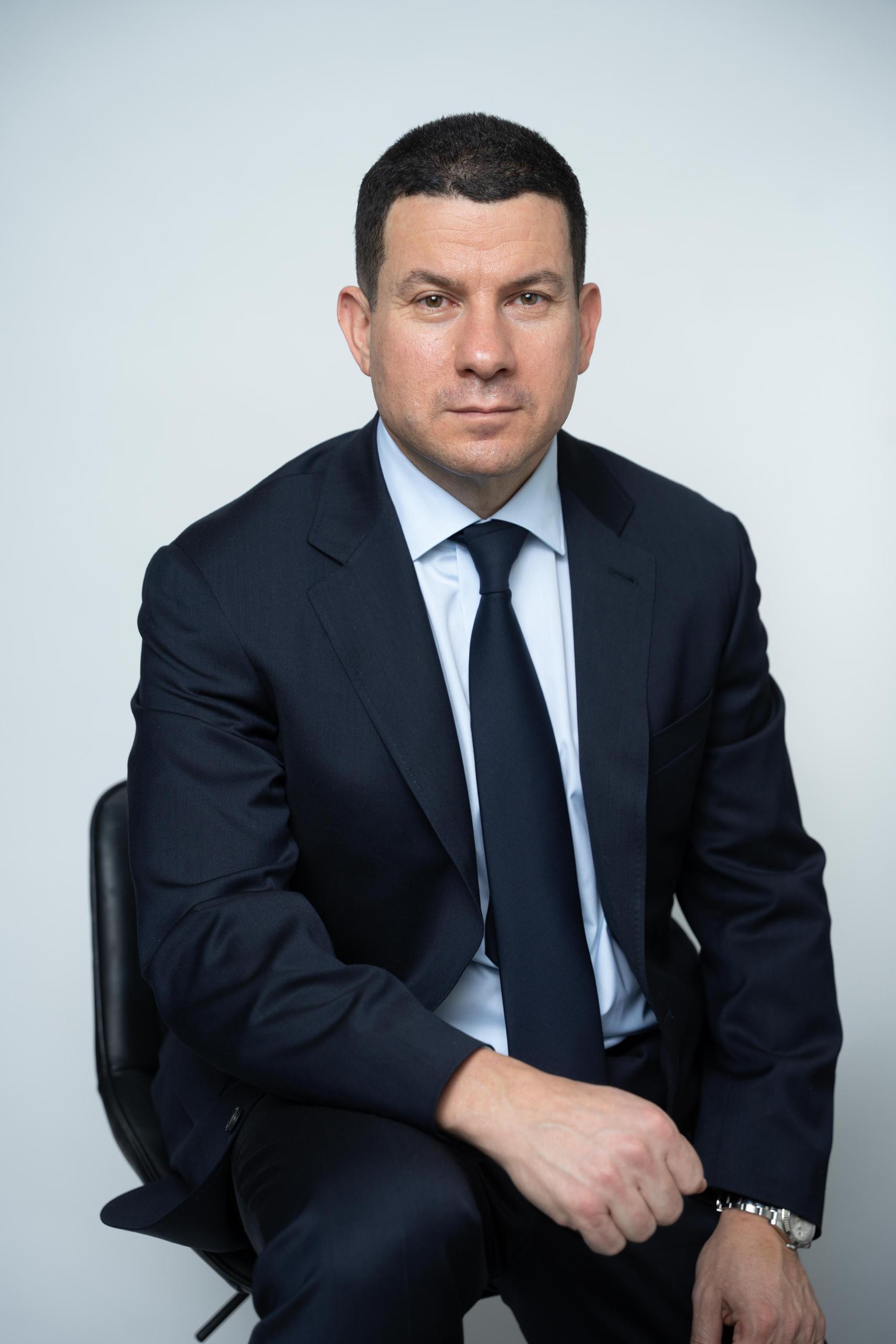 Sam wearing a blue suit and sitting on a chair smiling and looking at the camera.